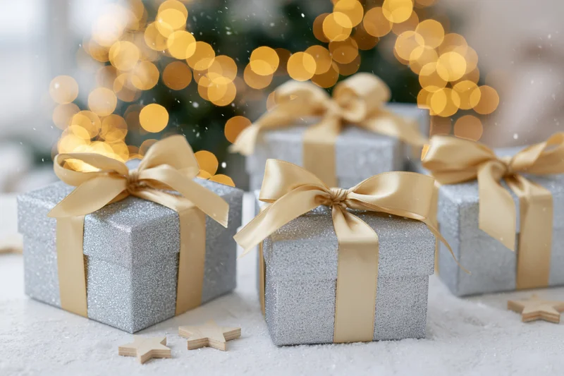 Silver glitter gift boxes with gold satin ribbon bows arranged on snowy surface near blurred christmas tree lights