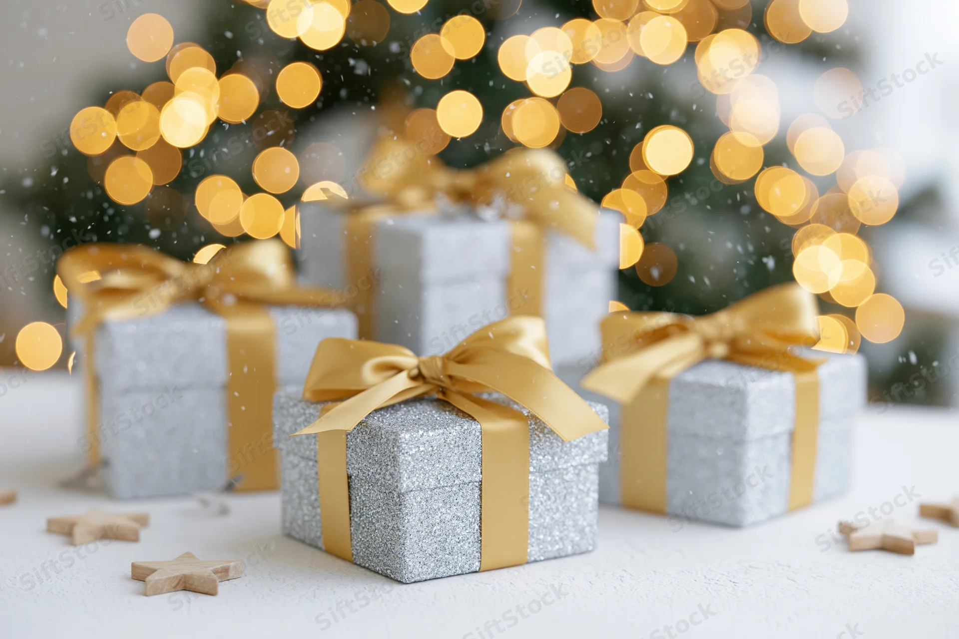 Silver glitter gift boxes with gold satin ribbon bows arranged on a white surface in front of blurred christmas lights and scattered wooden stars