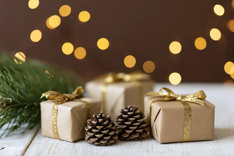 Small gift boxes wrapped in kraft paper with gold ribbon and pinecones on wooden table with blurred holiday lights in background
