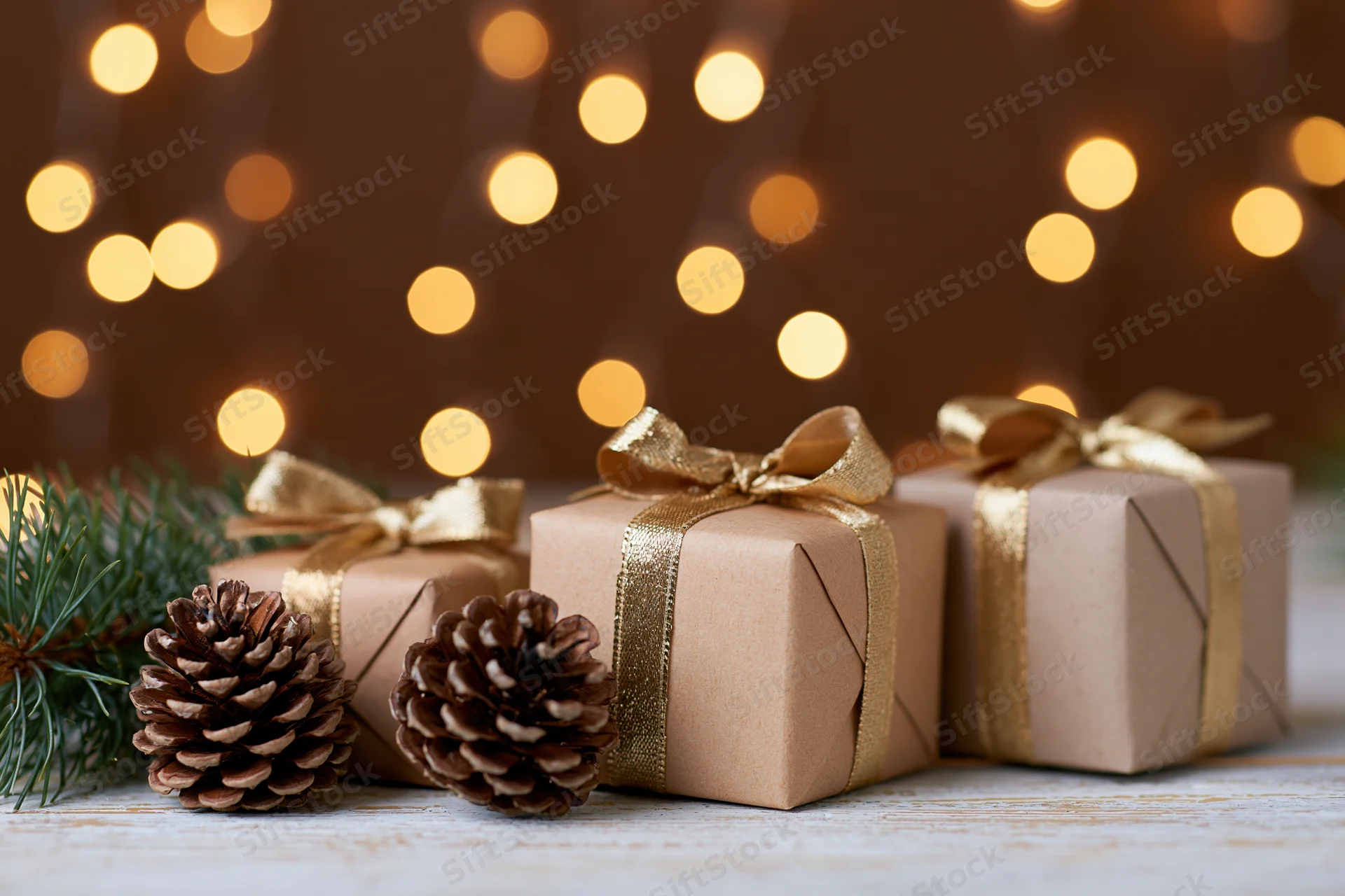 Three kraft-wrapped gift boxes with gold ribbons and pinecones on a wooden surface against warm bokeh lights