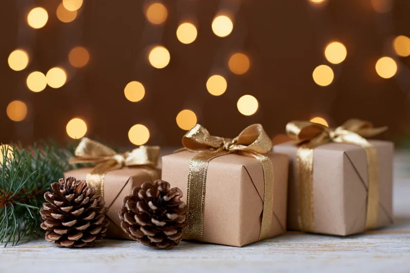 Three kraft-wrapped gift boxes with gold ribbons and pinecones on a wooden surface against warm bokeh lights