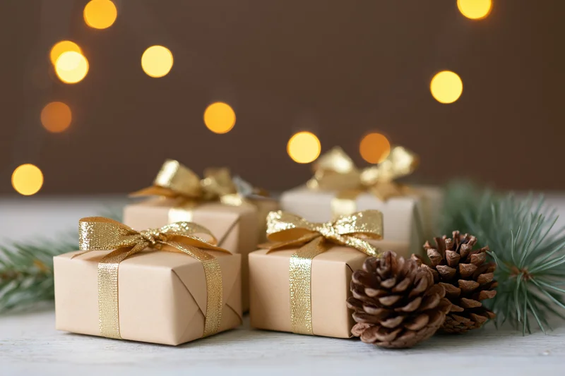 Small gift boxes wrapped in kraft paper with gold ribbons on a wooden surface with pinecones and blurred lights in the background