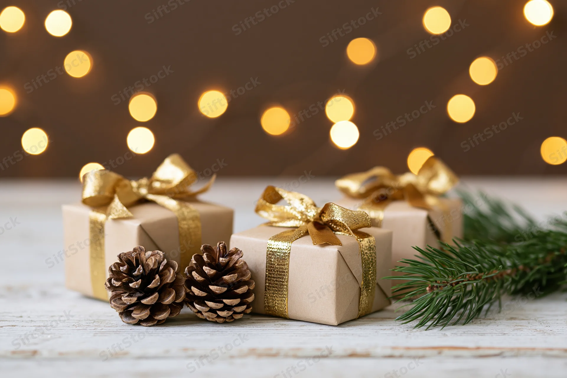 Three small gift boxes wrapped in kraft paper with gold ribbons on a wooden surface with pinecones and evergreen branches, warm bokeh lights in the background