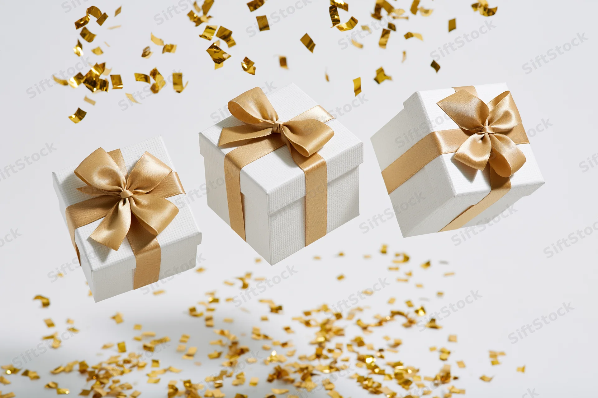 Three white gift boxes with gold satin ribbons and bows floating amidst falling golden confetti on a white background