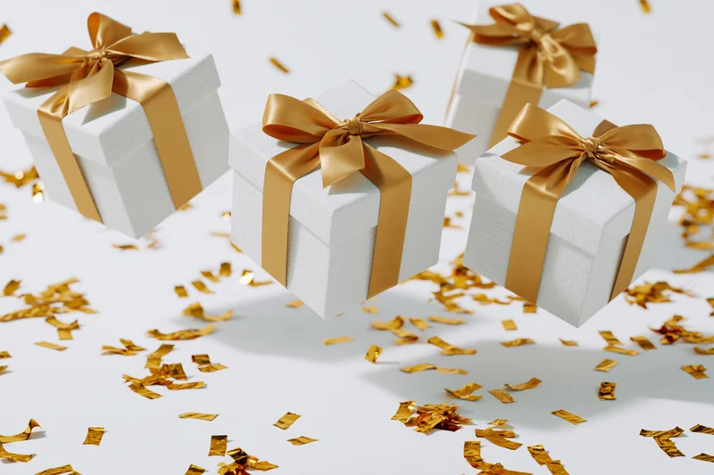 White gift boxes with gold satin ribbons and bows surrounded by scattered golden confetti on a white surface