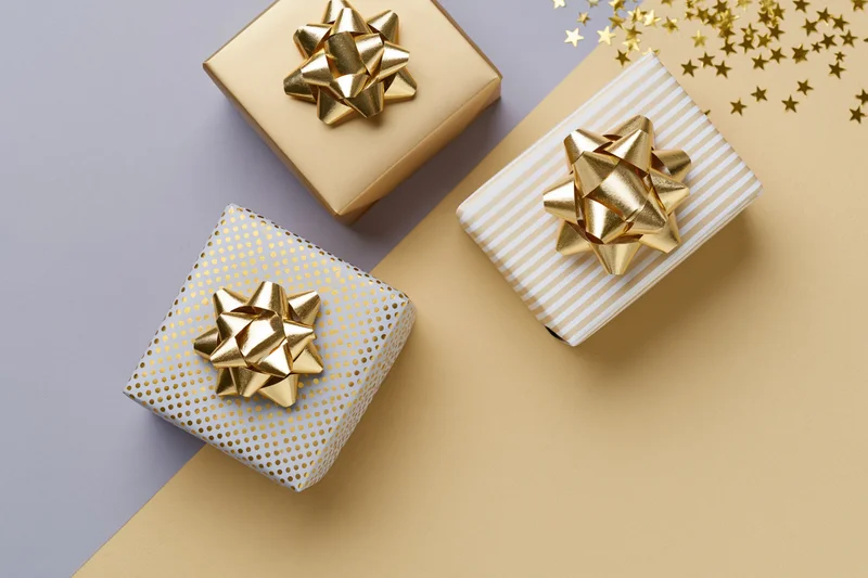 Three wrapped gift boxes with gold foil bows arranged diagonally on a two-tone surface with star confetti