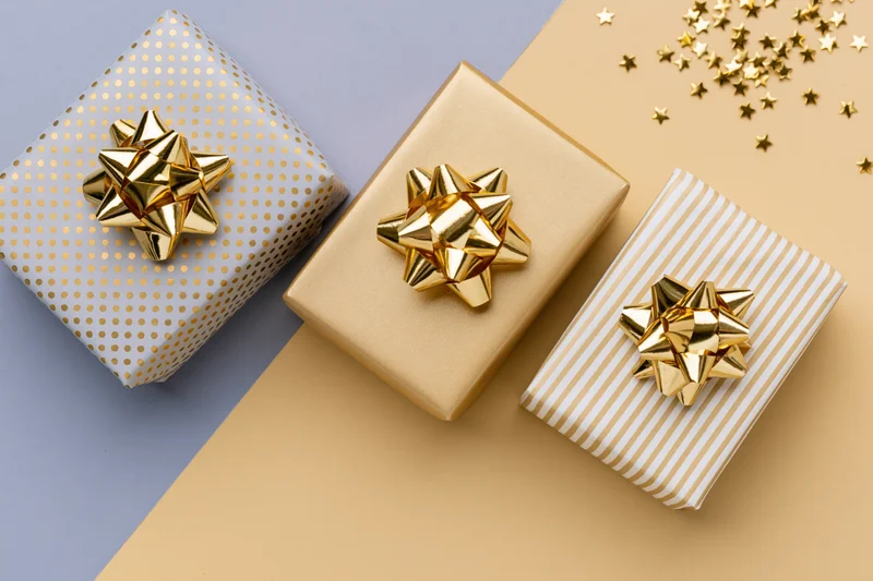 Three wrapped gift boxes with gold foil bows arranged diagonally on a two-tone background with scattered star confetti