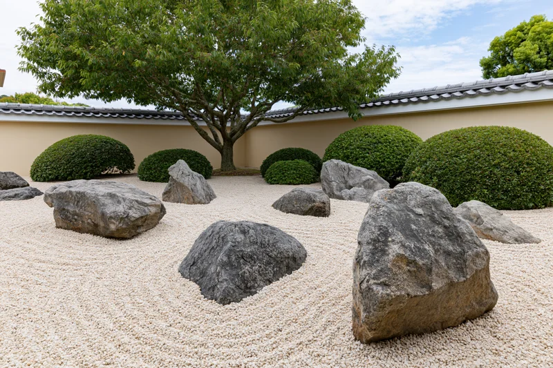 Traditional japanese zen garden with raked gravel, large rocks, and trimmed shrubs under a tree near a tiled roof wall