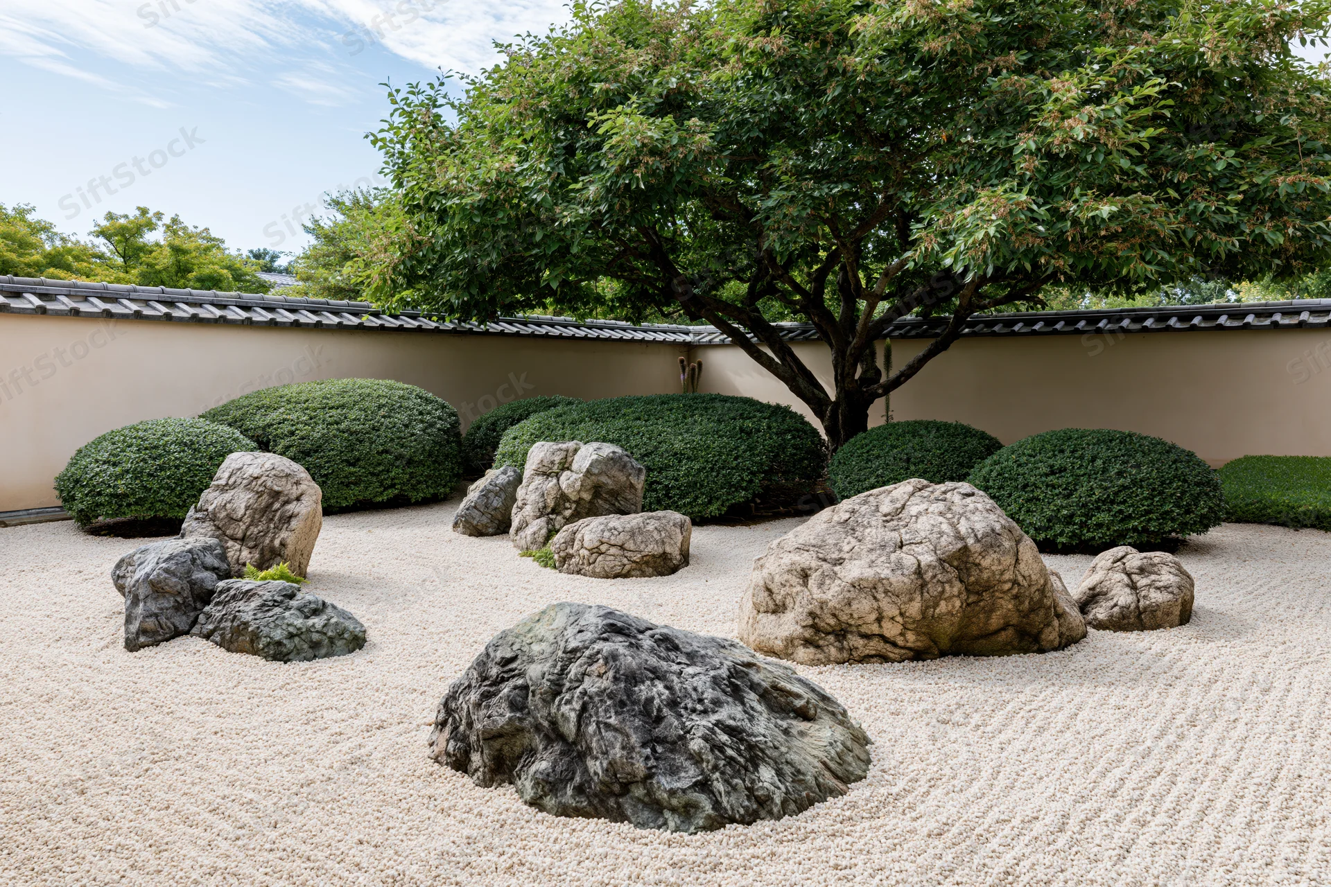 Raked gravel and arranged rocks in a traditional japanese zen garden surrounded by trimmed shrubs and a tree against a tiled wall