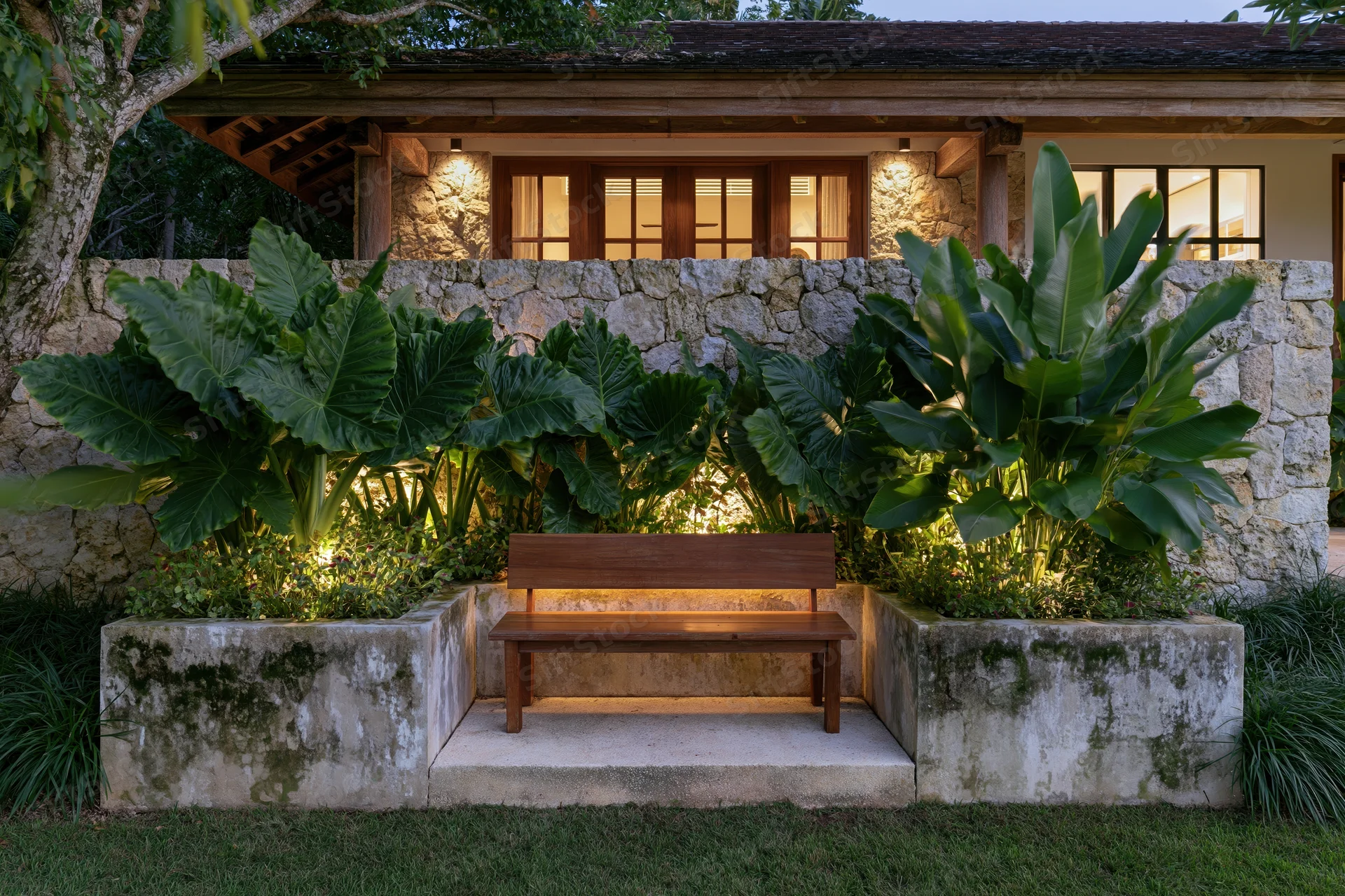 Wooden bench centered in a tropical garden courtyard at night, flanked by large leafy plants and stone walls, illuminated by warm ambient lighting