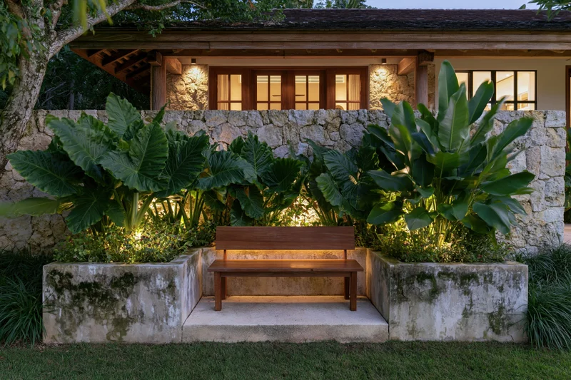 Wooden bench centered in a tropical garden courtyard at night, flanked by large leafy plants and stone walls, illuminated by warm ambient lighting