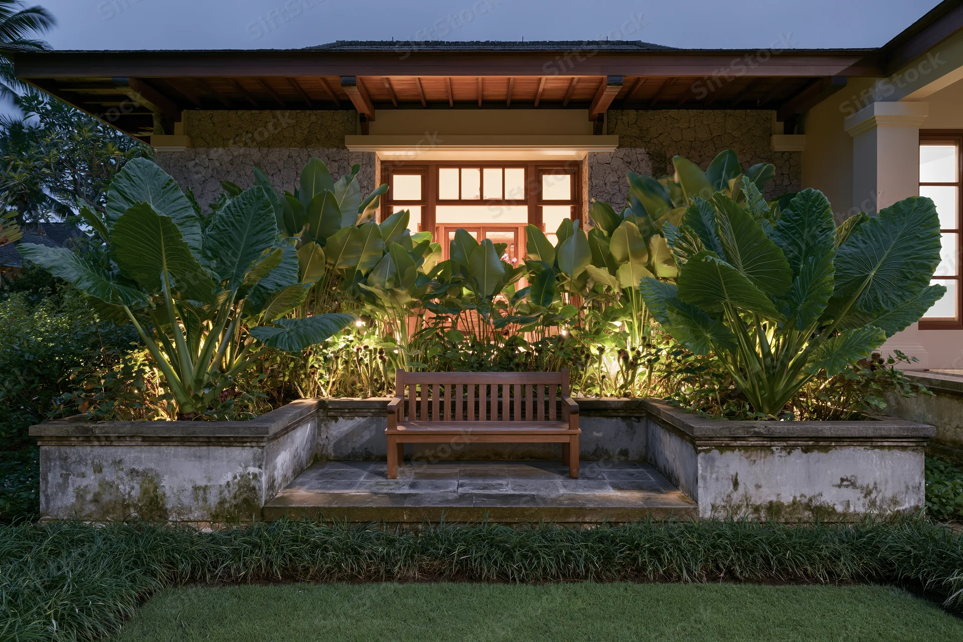 Wooden bench centered in a tropical garden courtyard at night, illuminated by warm lighting with large green plants and stone steps leading to a house porch