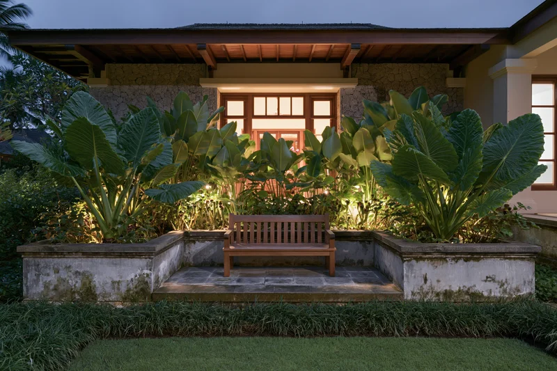 Wooden bench centered in a tropical garden courtyard at night, illuminated by warm lighting with large green plants and stone steps leading to a house porch