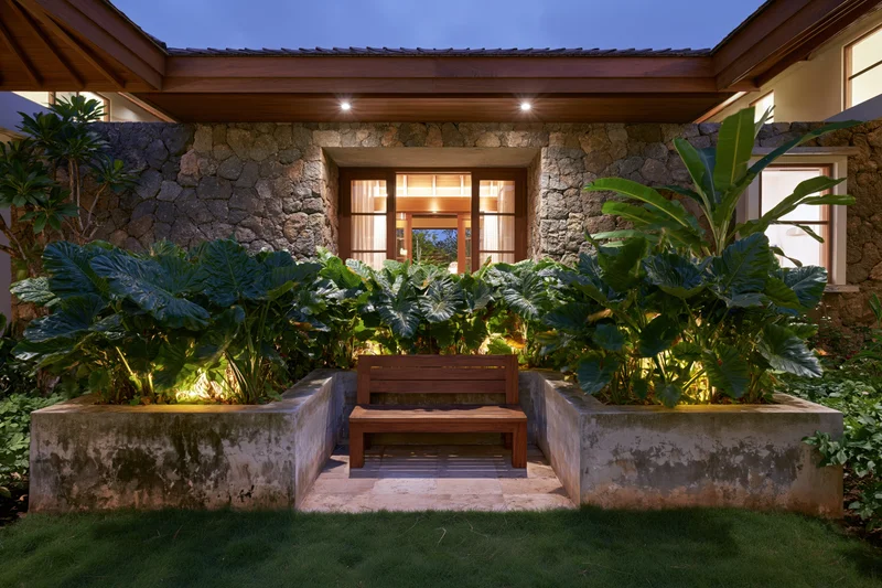 Wooden bench centered in a tropical garden courtyard at night, flanked by illuminated planters and stone walls