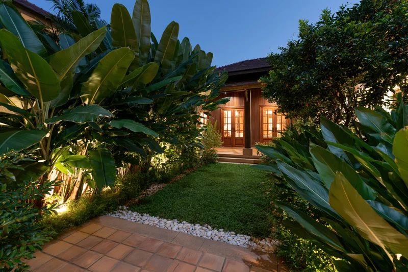 Tropical villa courtyard garden at night with illuminated banana plants and tiled pathway leading to wooden entrance