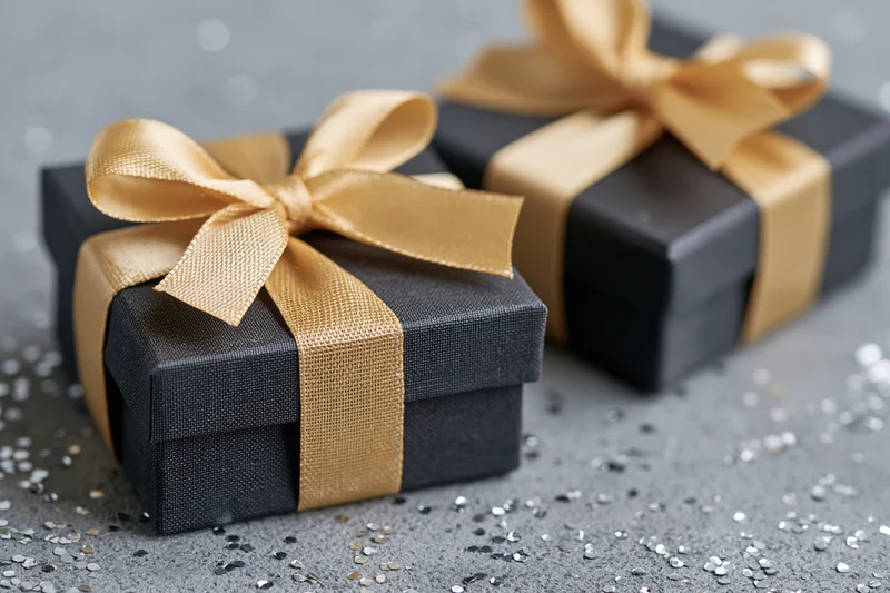 Two dark charcoal gift boxes tied with satin gold ribbon bows on a gray surface with scattered glitter