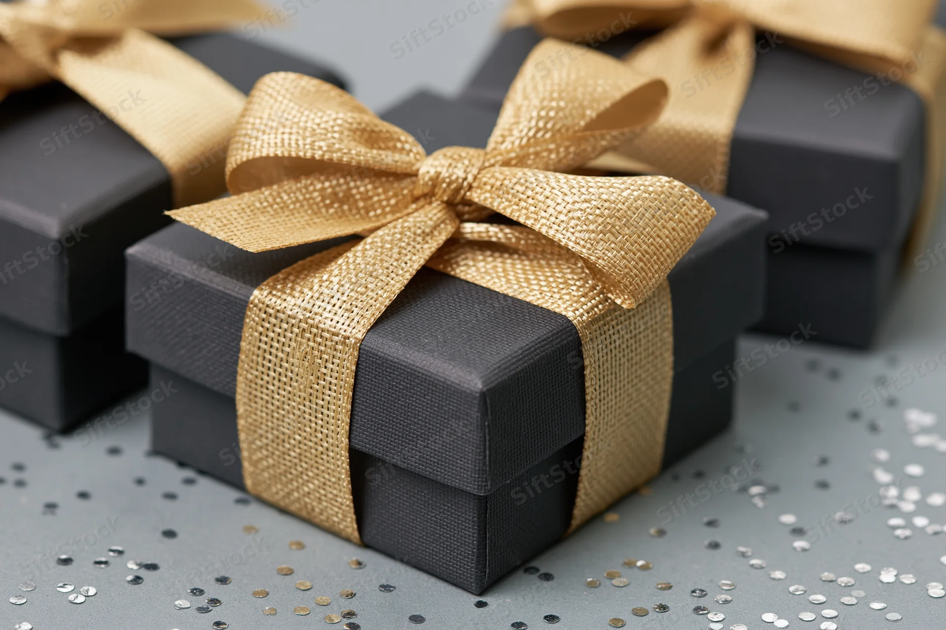 Dark charcoal gift boxes tied with gold ribbon bows on a surface with scattered confetti