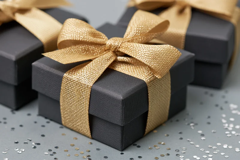 Dark charcoal gift boxes tied with gold ribbon bows on a surface with scattered confetti
