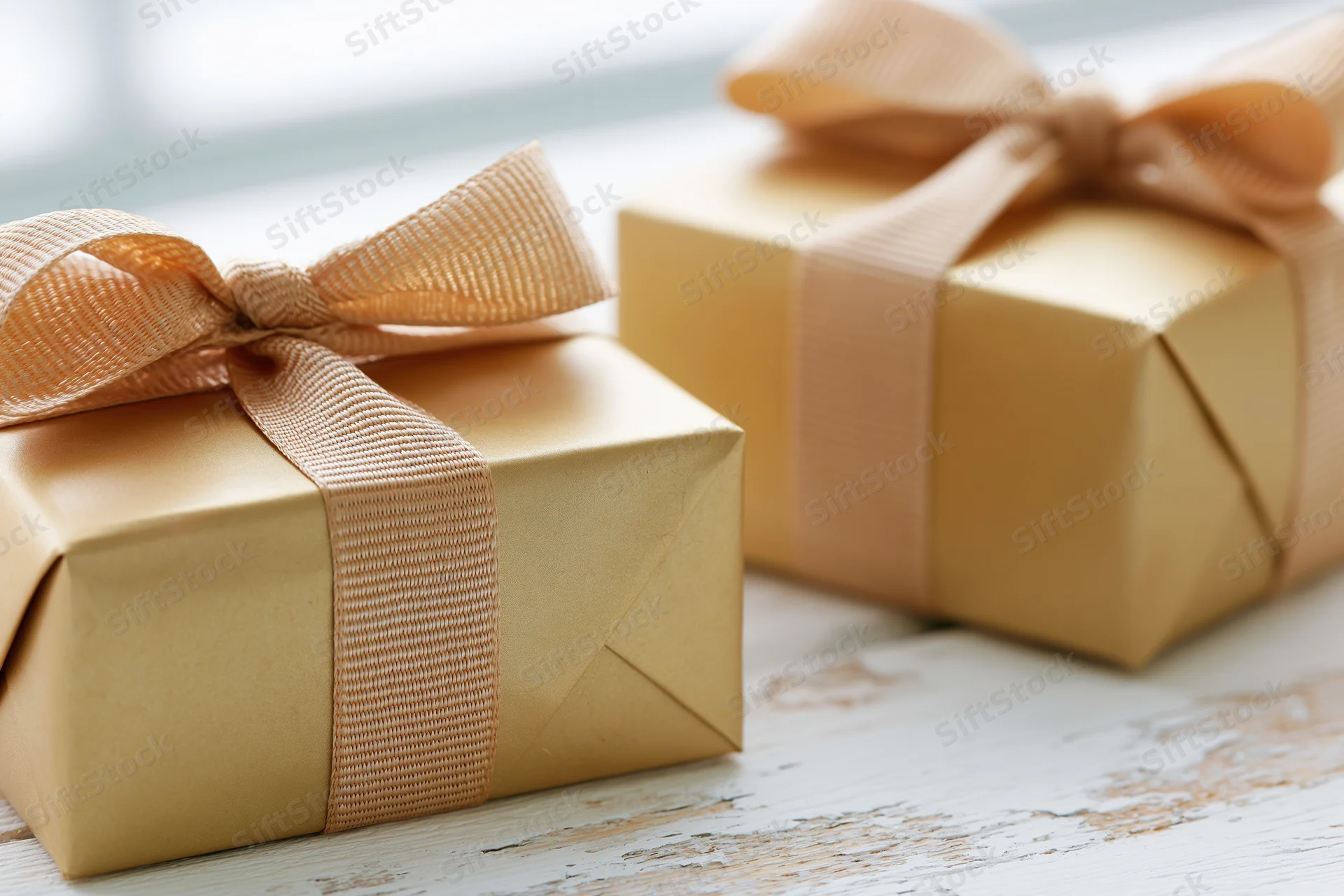 Two gold-wrapped gift boxes with peach ribbons on a rustic white wooden table