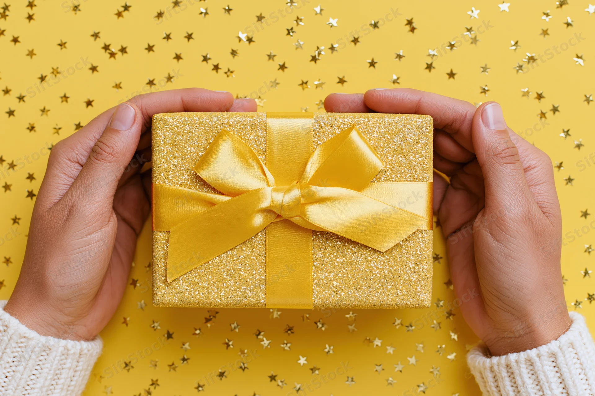 Hands holding a gold glitter gift box with yellow ribbon bow against starry yellow background