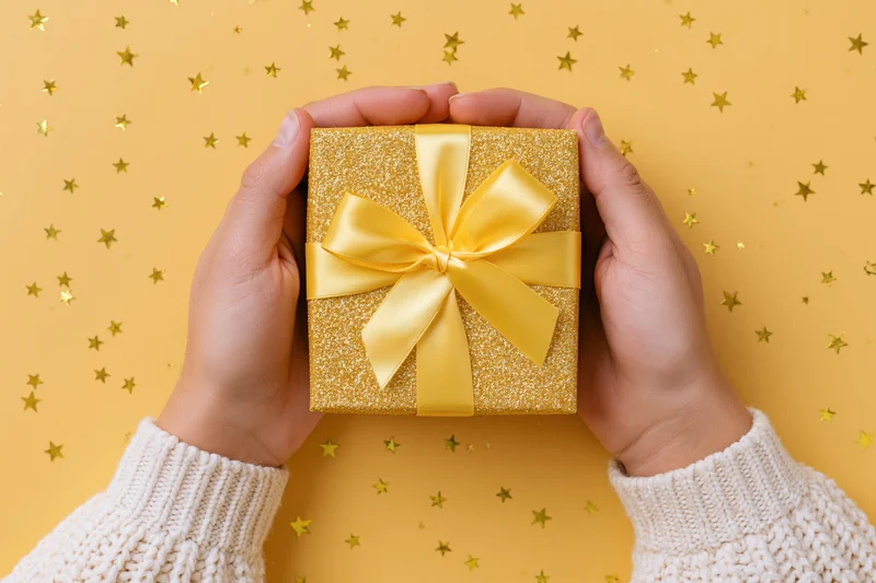 Hands holding a gold glitter gift box with yellow ribbon against a starry yellow background