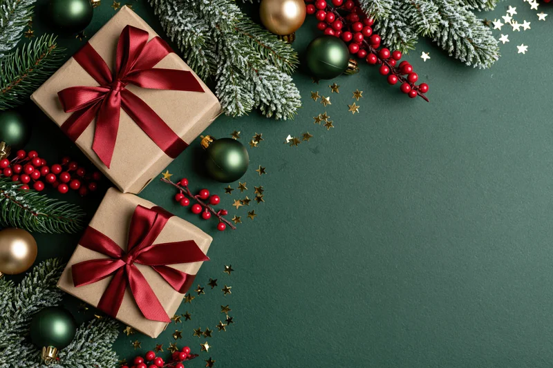 Two kraft paper gift boxes with red satin ribbon bows arranged with pine branches, ornaments, berries and stars on dark green surface