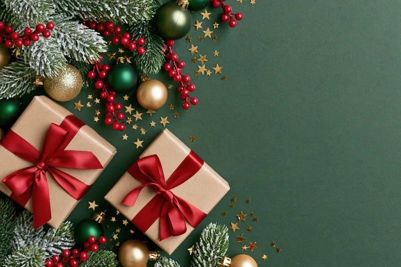 Two kraft paper gift boxes with red satin ribbon bows arranged with snowy pine branches, red berries, gold stars and ornaments on dark green background