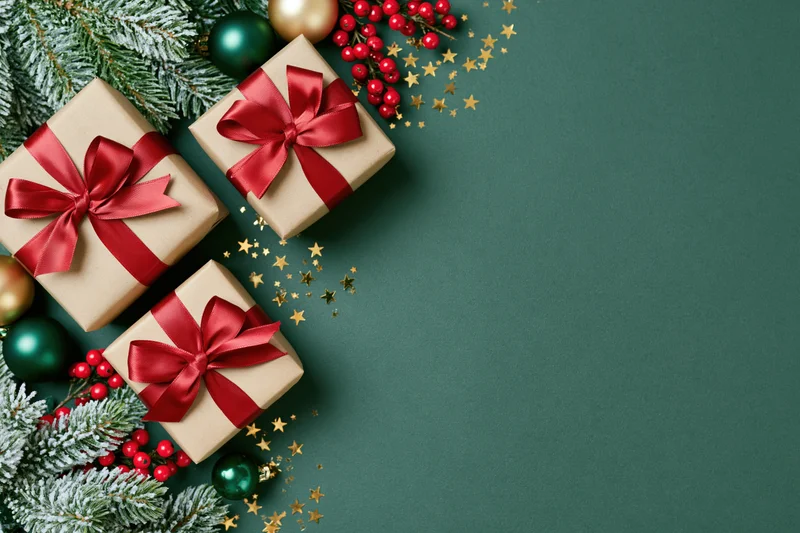 Three kraft paper gift boxes with red satin ribbon bows arranged with pine branches, berries, ornaments and gold stars on a green surface