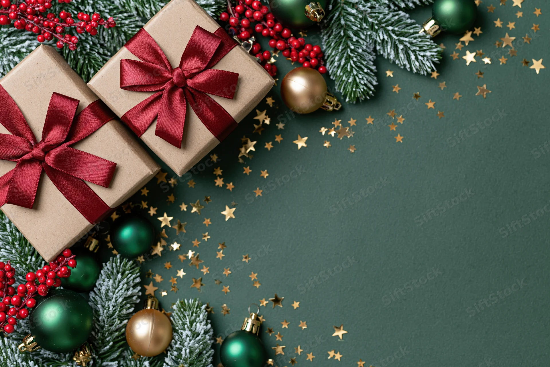 Two kraft paper gift boxes with red satin ribbon bows arranged with pine branches, berries, ornaments and gold stars on dark green surface