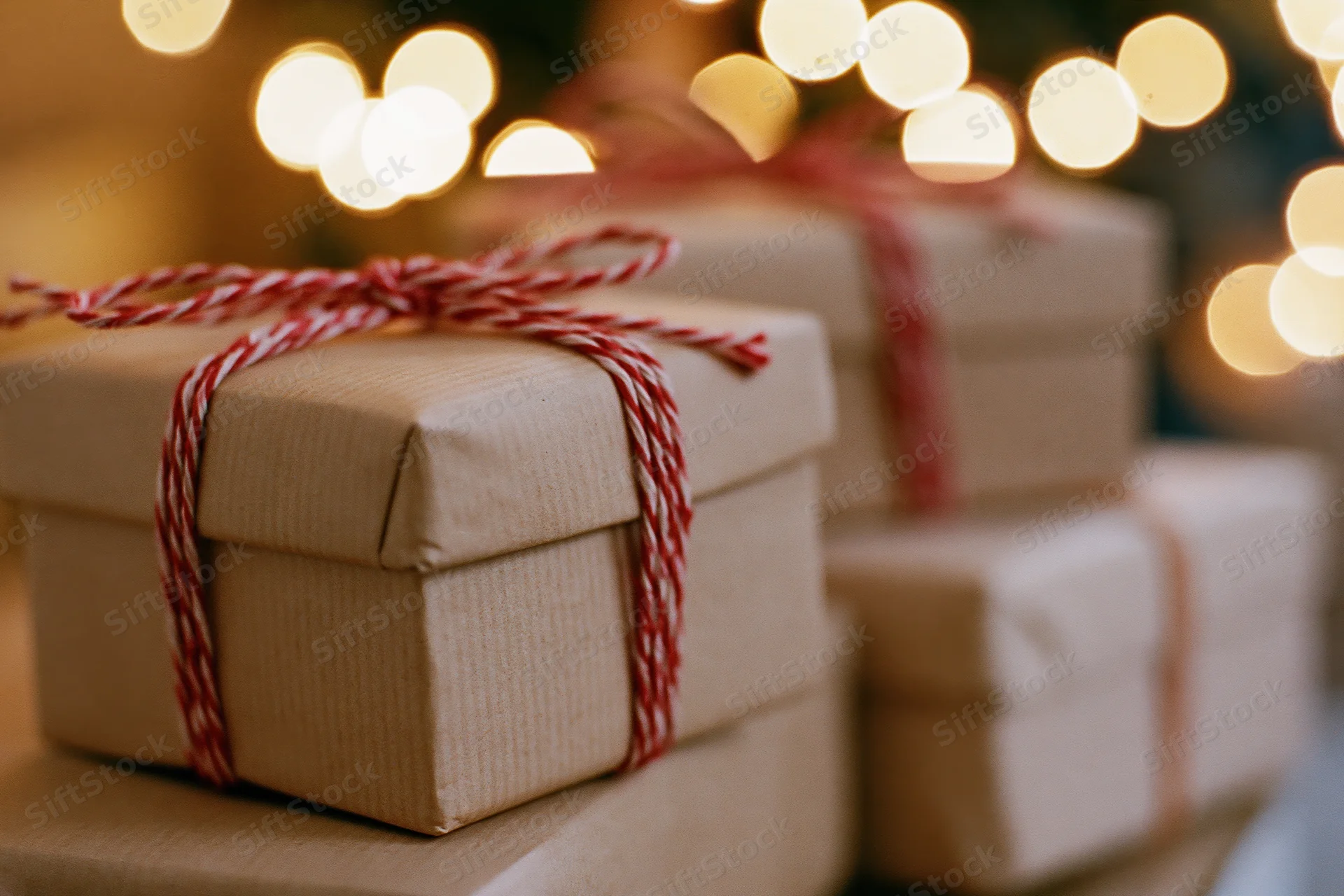 Stacked kraft paper gift boxes tied with red and white twine bows in front of blurred holiday lights