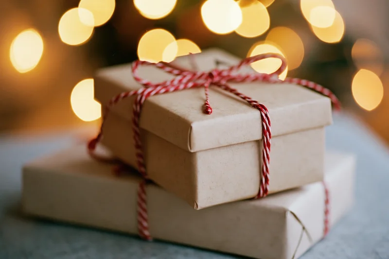 Two kraft paper gift boxes stacked and tied with red twine bows on a soft surface with warm bokeh lights in the background
