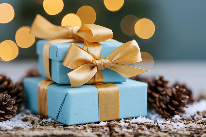 Two light blue gift boxes tied with gold satin ribbons stacked on snowy wood with pinecones and bokeh lights behind
