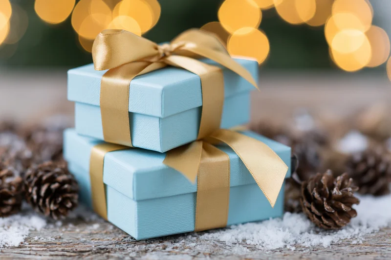 Two light blue gift boxes tied with gold satin ribbons stacked on snowy wooden surface with pinecones and blurred holiday lights in background