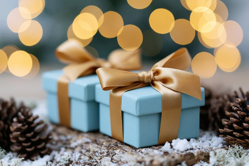 Two light blue gift boxes with gold satin ribbons on snowy wood surrounded by pinecones and bokeh lights