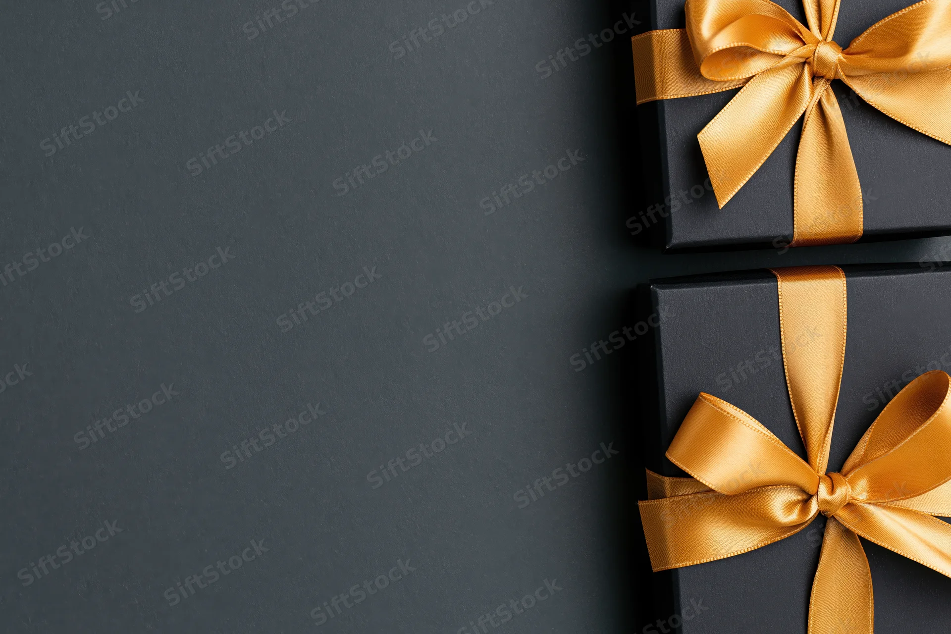 Two matte black gift boxes with gold satin ribbon bows on dark surface