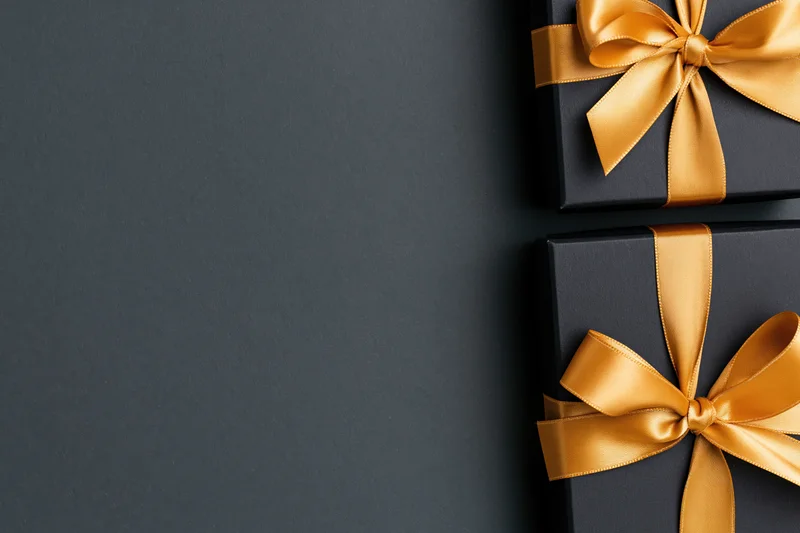 Two matte black gift boxes with gold satin ribbon bows on dark surface