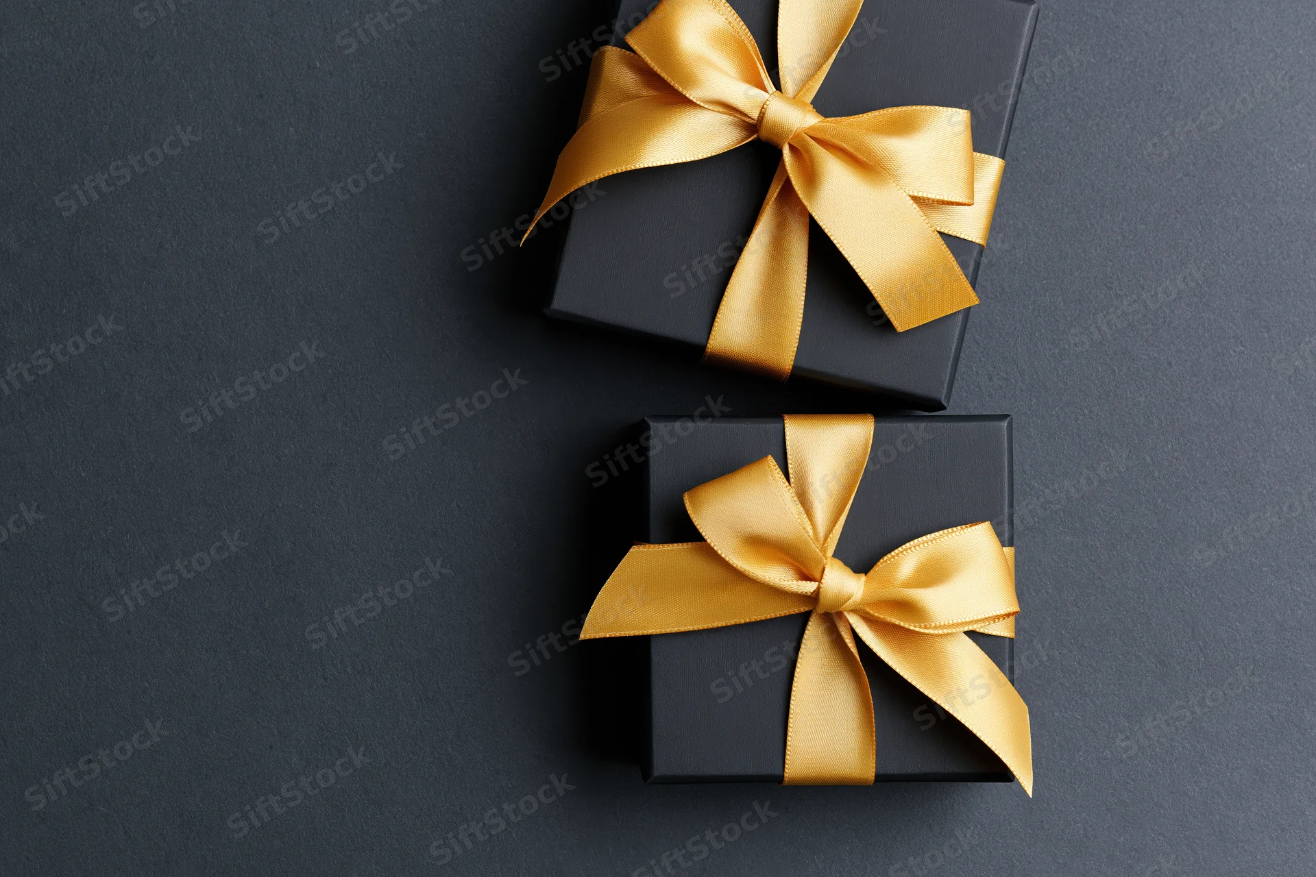 Two matte black gift boxes with gold satin ribbon bows on dark surface