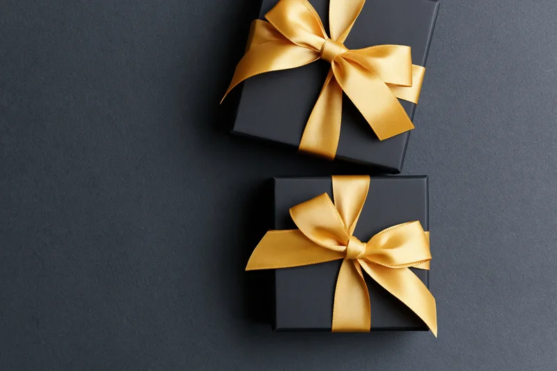 Two matte black gift boxes with gold satin ribbon bows on dark surface