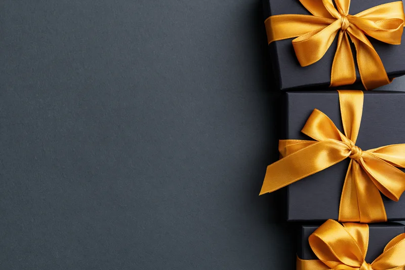 Three matte black gift boxes with gold satin ribbon bows arranged vertically on dark surface with copyspace on left