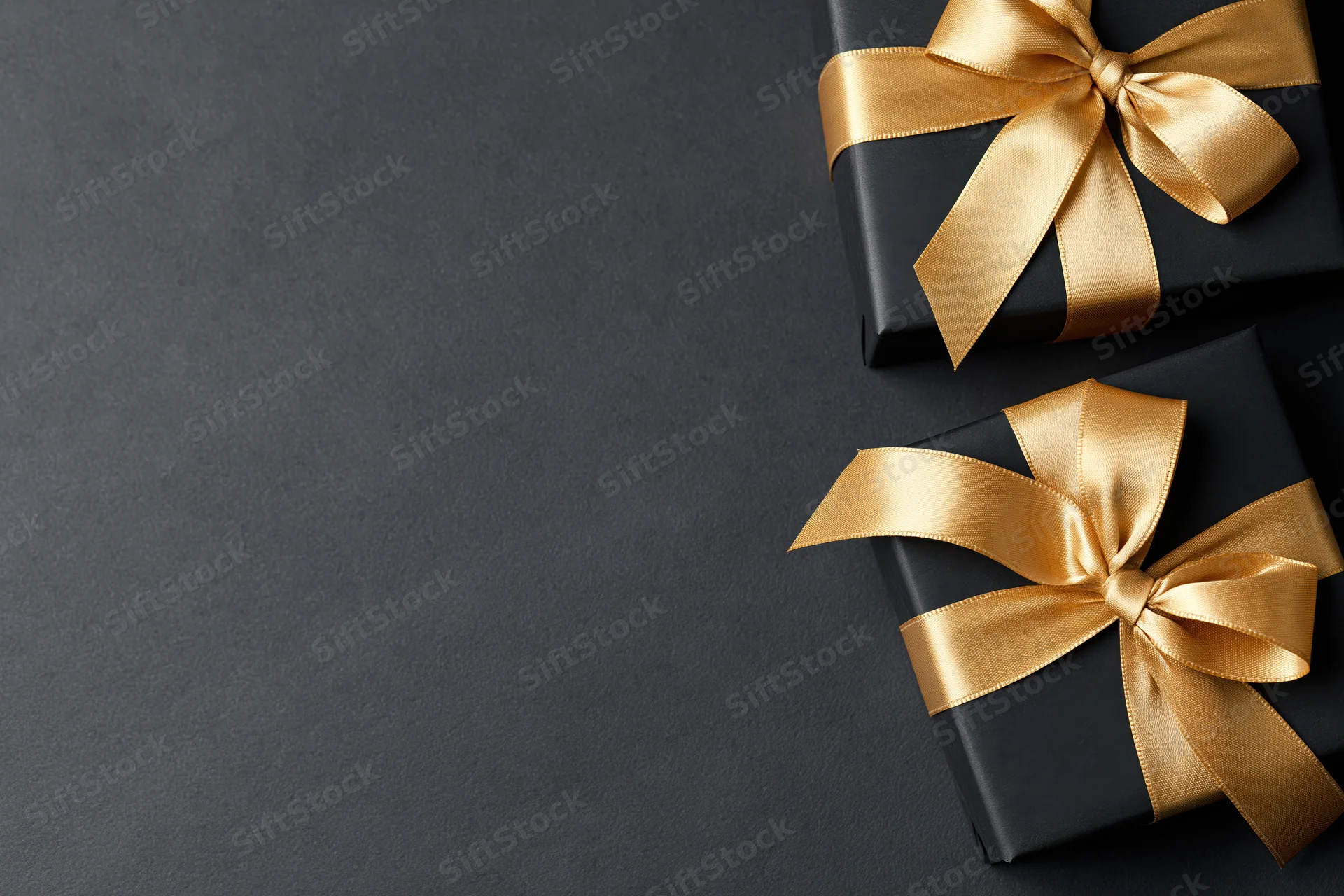 Two matte black gift boxes with gold satin ribbon bows on dark surface