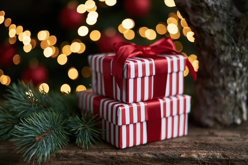 Two red and white striped gift boxes stacked on a wooden surface near a decorated christmas tree with bokeh lights