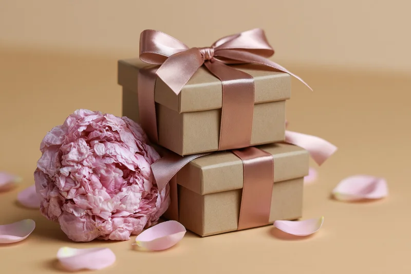 Two stacked kraft gift boxes with pink ribbon and a peony flower beside them on a beige surface