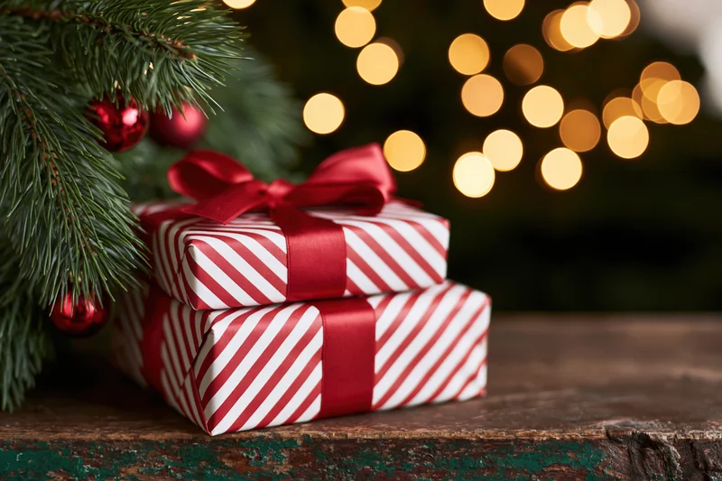 Two stacked gift boxes wrapped in red and white diagonal stripes with red ribbon and bow on a rustic wooden table beside a decorated christmas tree branch with blurred lights in the background