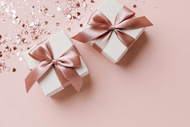 Two white gift boxes with dusty rose satin ribbon bows on pink surface with glitter and hearts