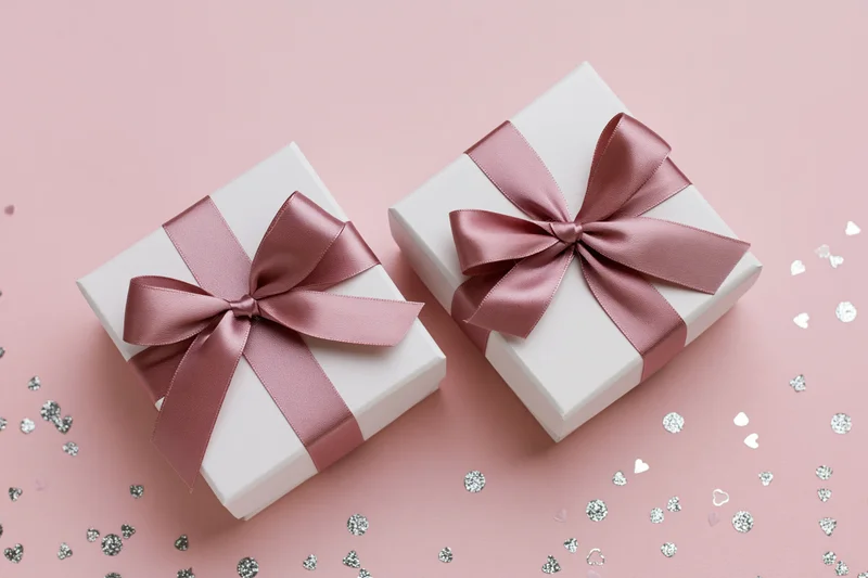 Two white gift boxes with dusty rose satin ribbon bows on pink surface with silver heart confetti