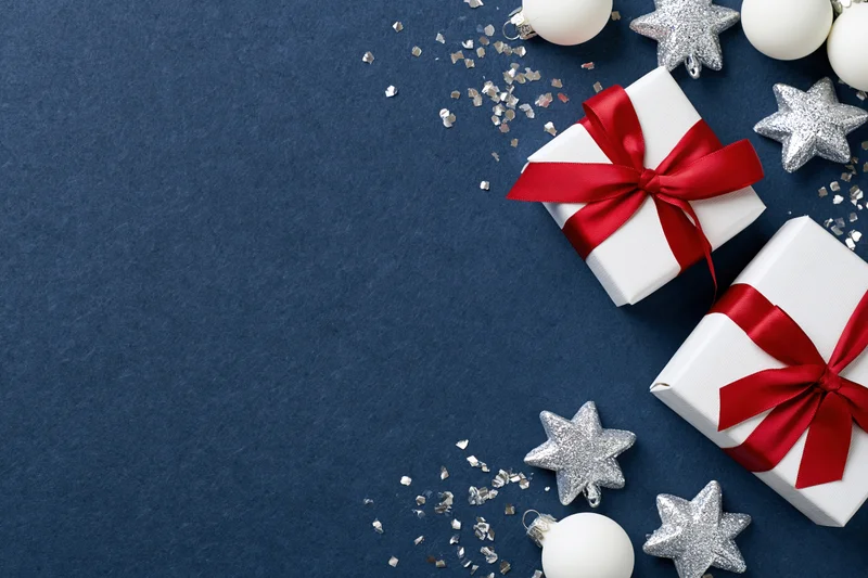 Two white gift boxes with red ribbons and bows on blue background with silver stars and confetti