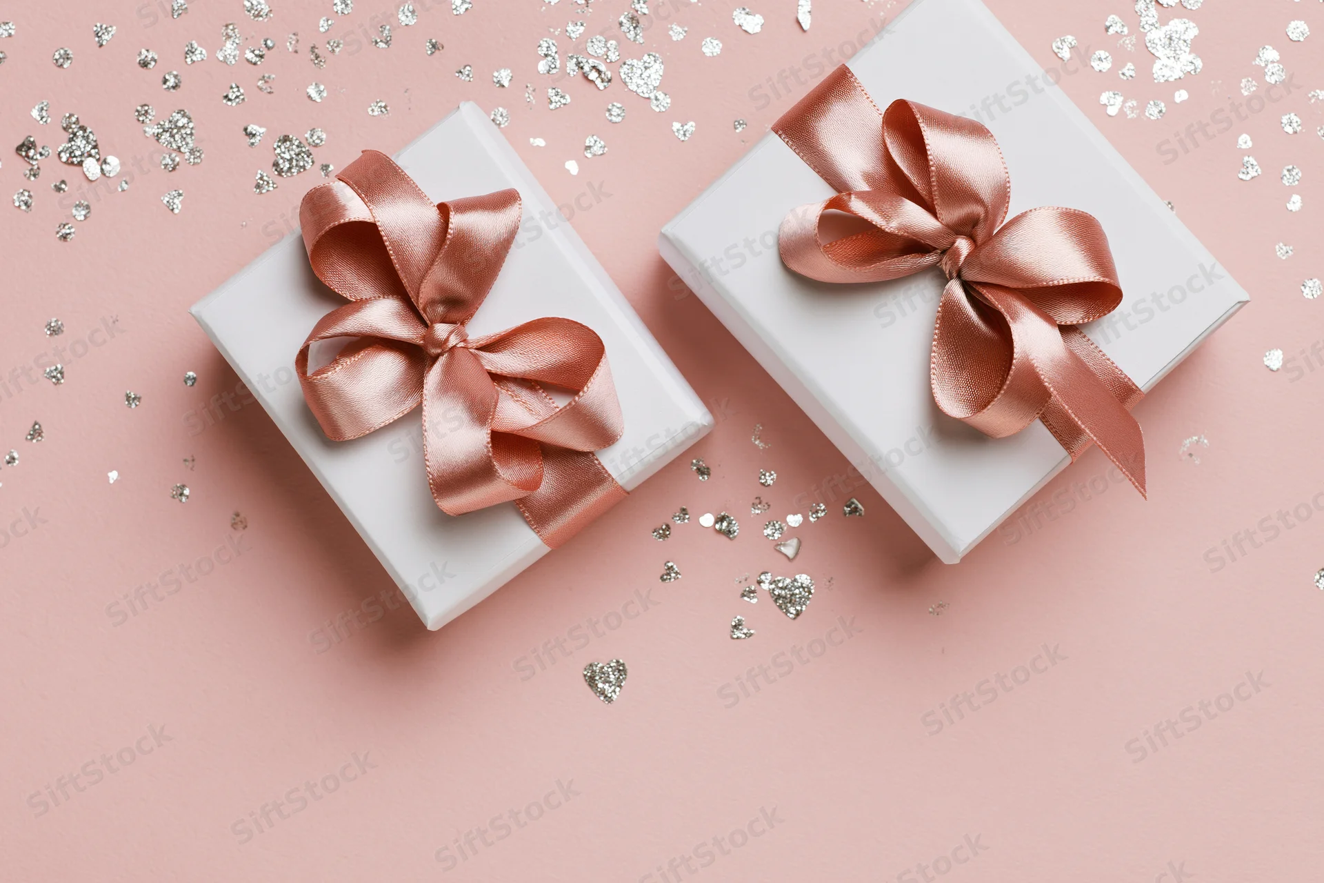 Two white gift boxes with rose-gold satin ribbon bows on pink surface with silver heart confetti