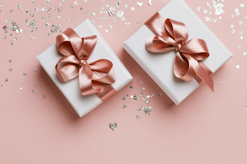 Two white gift boxes with rose-gold satin ribbon bows on pink surface with silver heart confetti