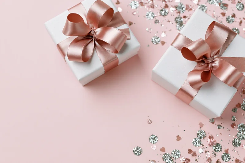 Two white gift boxes with rose-gold satin ribbon bows on pink surface with scattered confetti