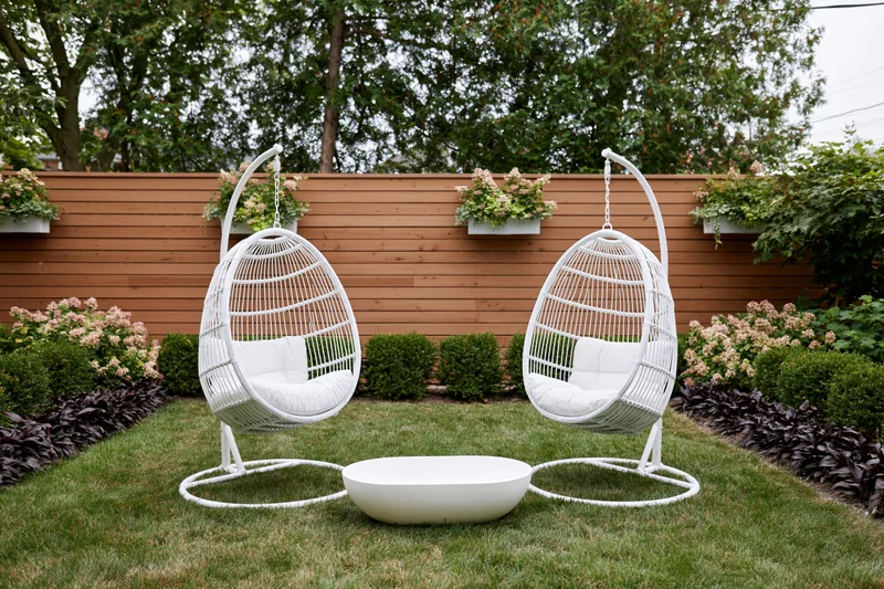 Two white rattan hanging egg chairs on freestanding frames in a grassy backyard garden with flowering shrubs and a wooden fence
